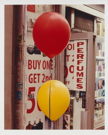 Two balloons, red and yellow, are tethered outside a storefront with advertisements and perfume on display in the windows.