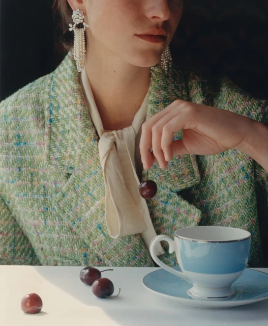 Woman in a green tweed blazer holds a cherry near a light blue teacup, with pearl earrings and a neutral-toned neck scarf.