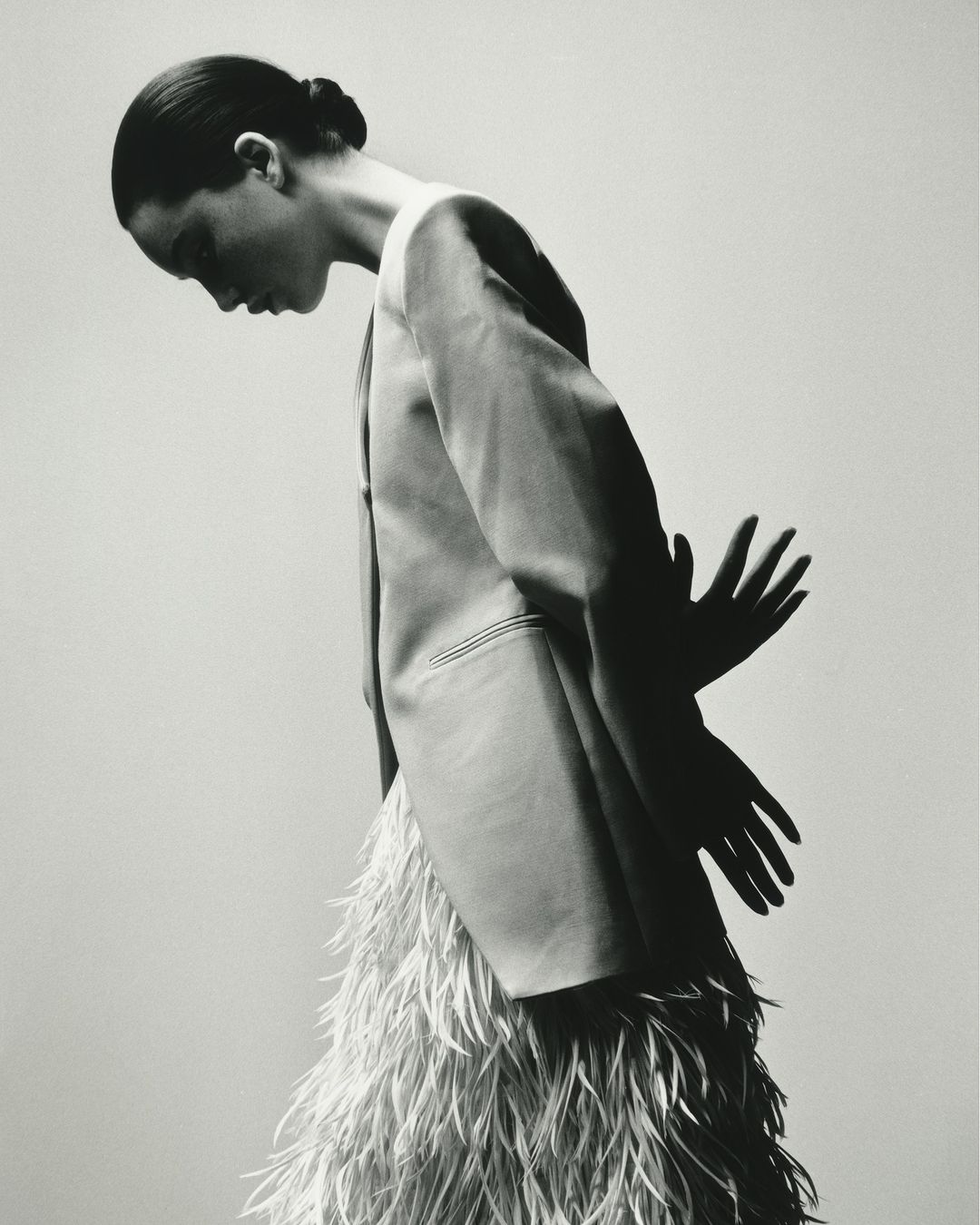 Black and white fashion shot: A woman in a blazer and feather skirt stands against a gray backdrop with a pair of shadow hands.