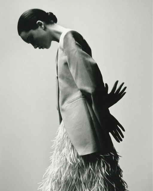 Black and white fashion shot: A woman in a blazer and feather skirt stands against a gray backdrop with a pair of shadow hands.