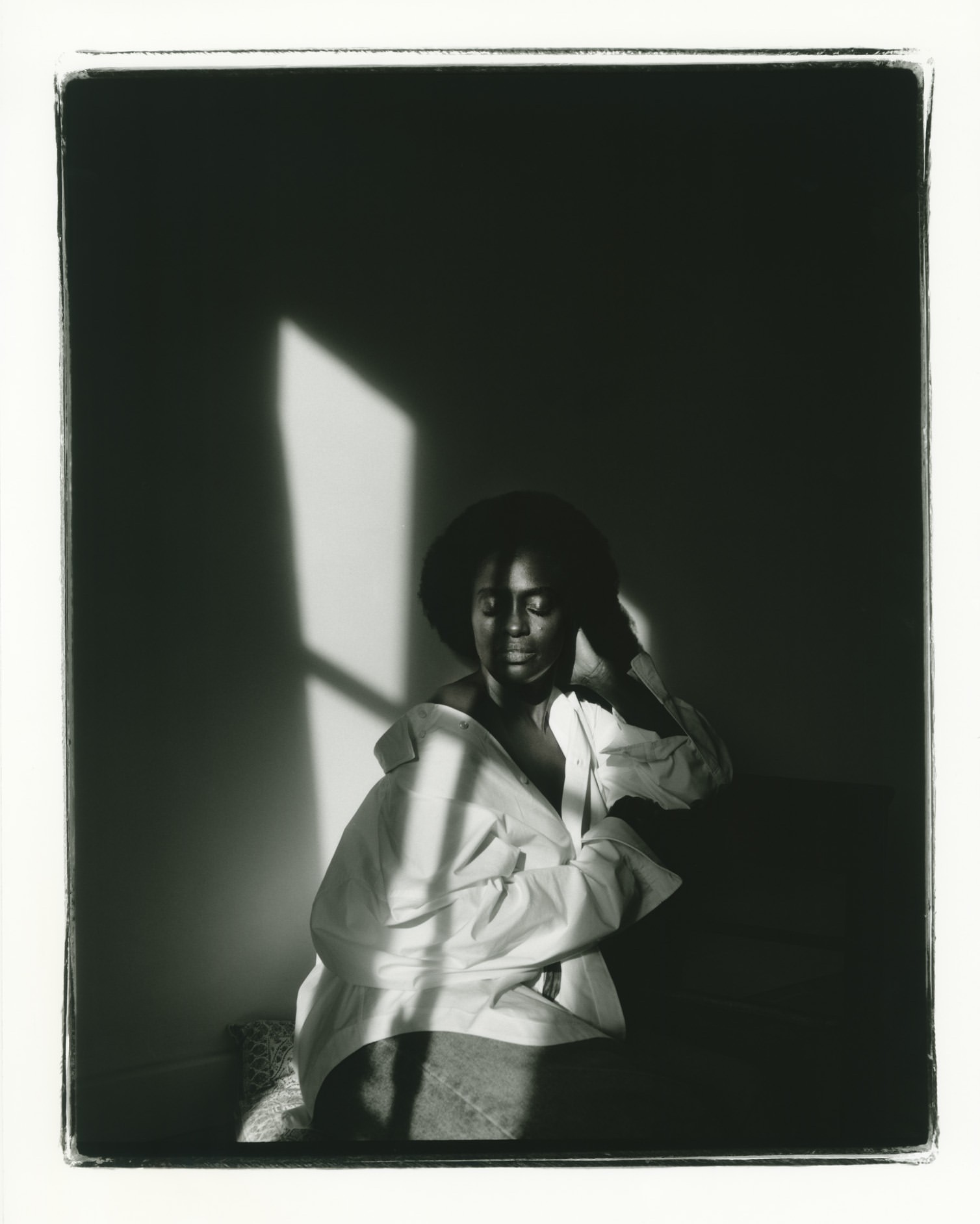 Black and white portrait of a woman with dark hair wearing an unbuttoned white shirt sitting indoors near a window with soft lighting.