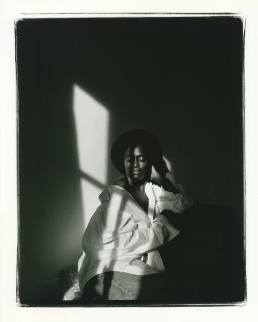 Black and white portrait of a woman with dark hair wearing an unbuttoned white shirt sitting indoors near a window with soft lighting.