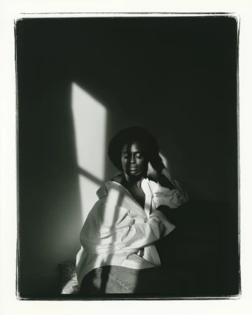 Black and white portrait of a woman with dark hair wearing an unbuttoned white shirt sitting indoors near a window with soft lighting.