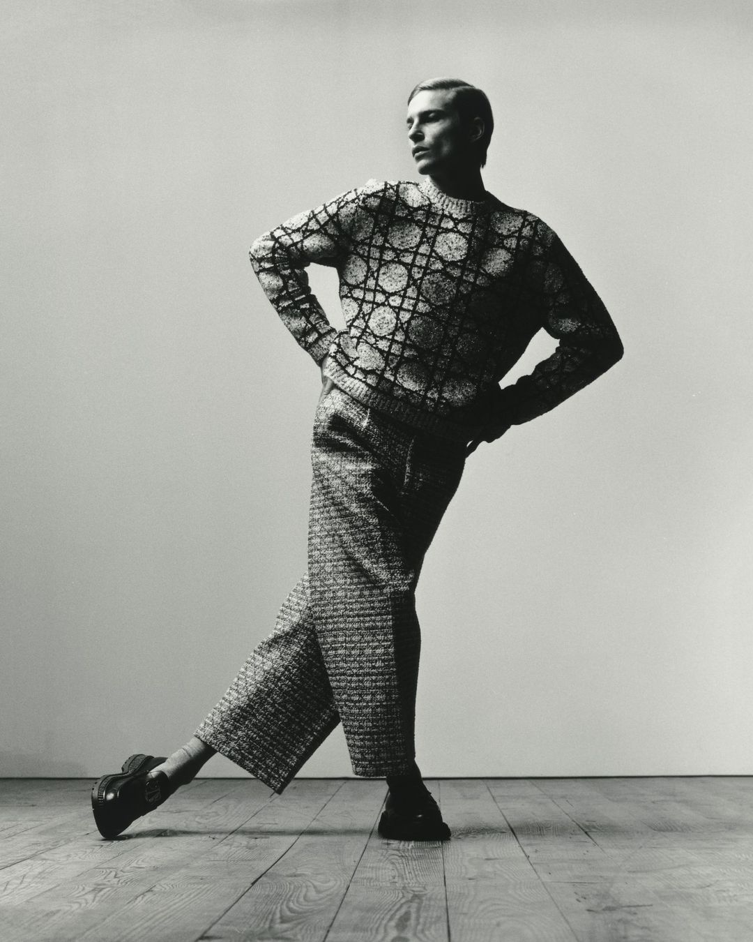 Black and white studio shot of adult male fashion model in patterned knit sweater and wide-legged pants striking a pose on wood floor.