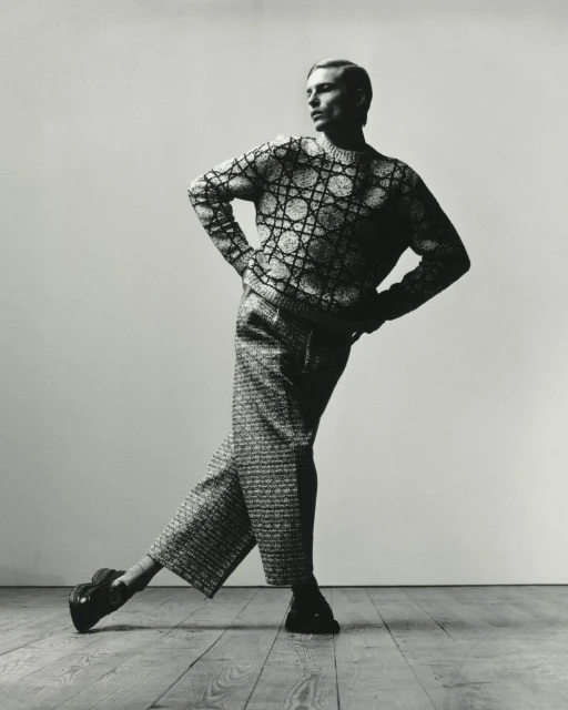 Black and white studio shot of adult male fashion model in patterned knit sweater and wide-legged pants striking a pose on wood floor.