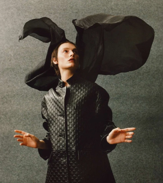 Fashion portrait of a young adult woman with short brown hair, wearing a textured black coat and a dramatic black headpiece against gray.