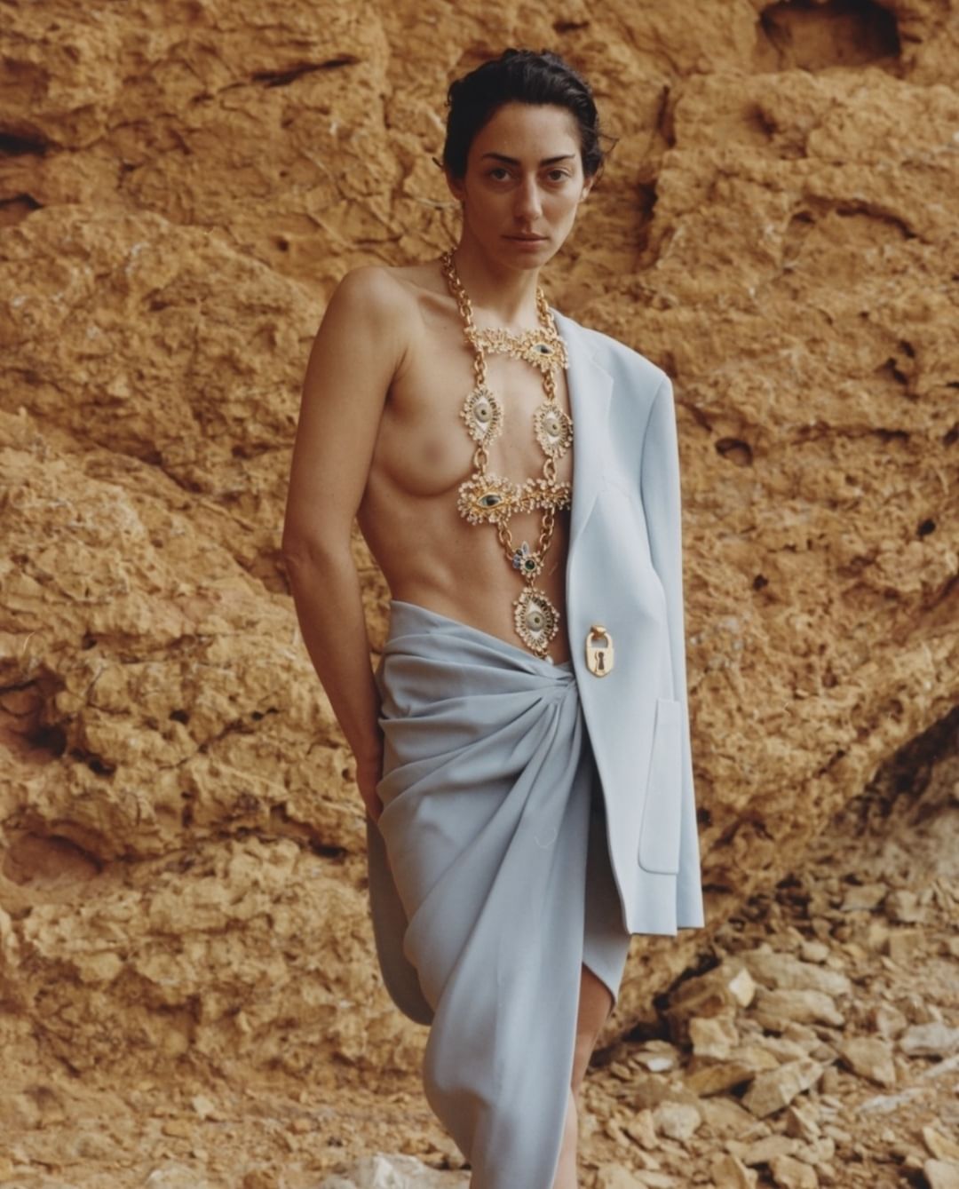Woman wearing gold necklace poses in front of a rock wall. She is wearing a light blue blazer draped on her shoulder.