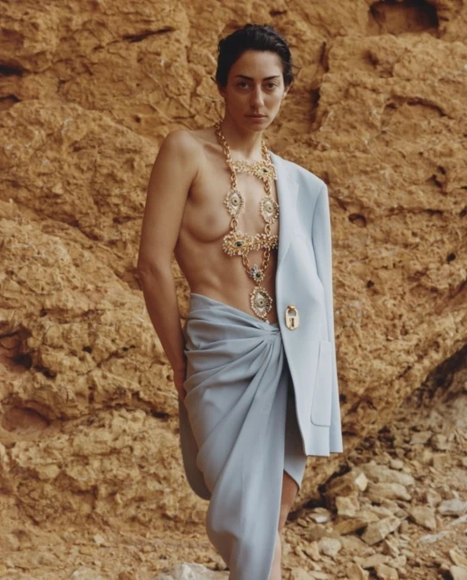 Woman wearing gold necklace poses in front of a rock wall. She is wearing a light blue blazer draped on her shoulder.
