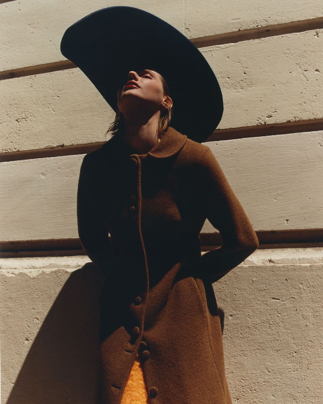 A woman in a brown coat and black wide-brimmed hat stands against a wall bathed in sunlight, fashion photography, outdoor setting.