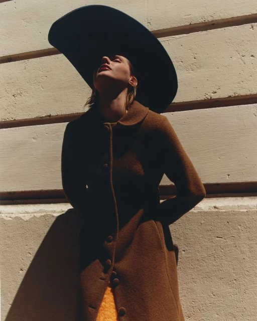 A woman in a brown coat and black wide-brimmed hat stands against a wall bathed in sunlight, fashion photography, outdoor setting.