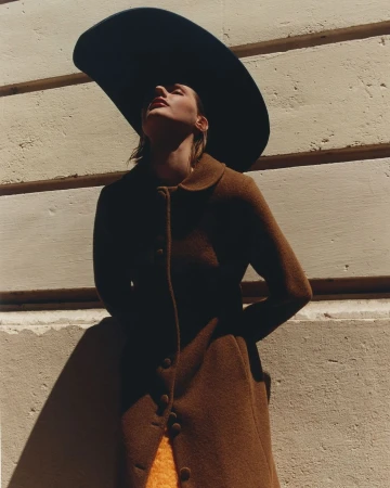 A woman in a brown coat and black wide-brimmed hat stands against a wall bathed in sunlight, fashion photography, outdoor setting.