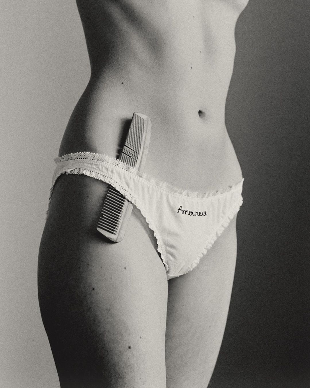 Monochrome close-up of a slender figure wearing underwear with a comb tucked into the waistband against a neutral studio backdrop.