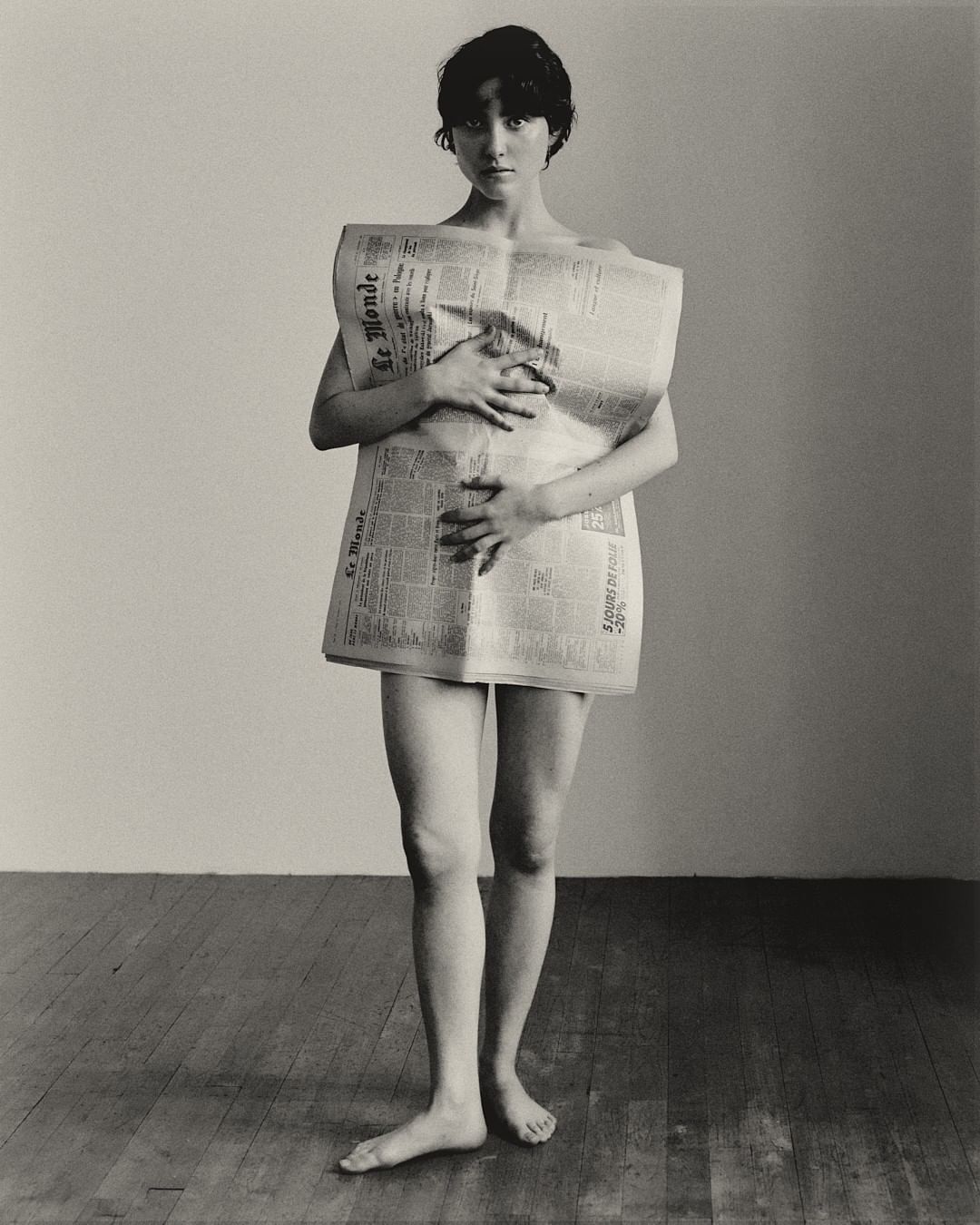 Striking black and white studio shot of a young adult woman wearing a newspaper as clothing, standing barefoot on a wood floor.