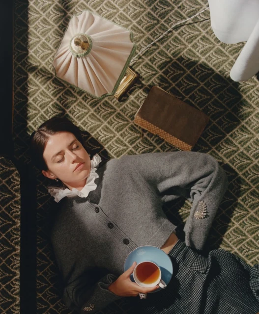Woman in gray cardigan lying on patterned rug with teacup, purse, and book. Interior shot, diffused light. Editorial lifestyle image.