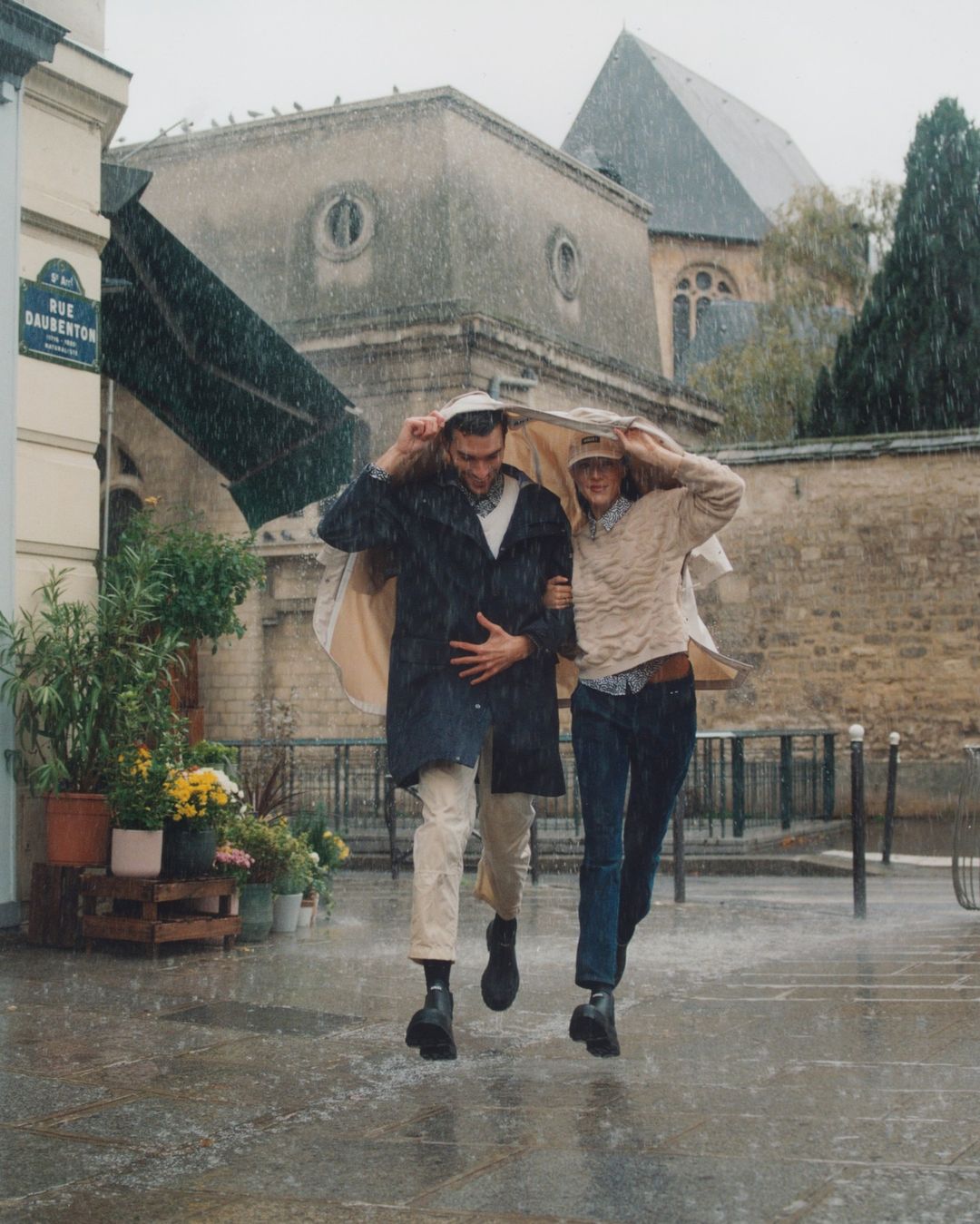 A couple walks together in heavy rain, using a jacket to shield themselves from the downpour, with a cityscape in the background.