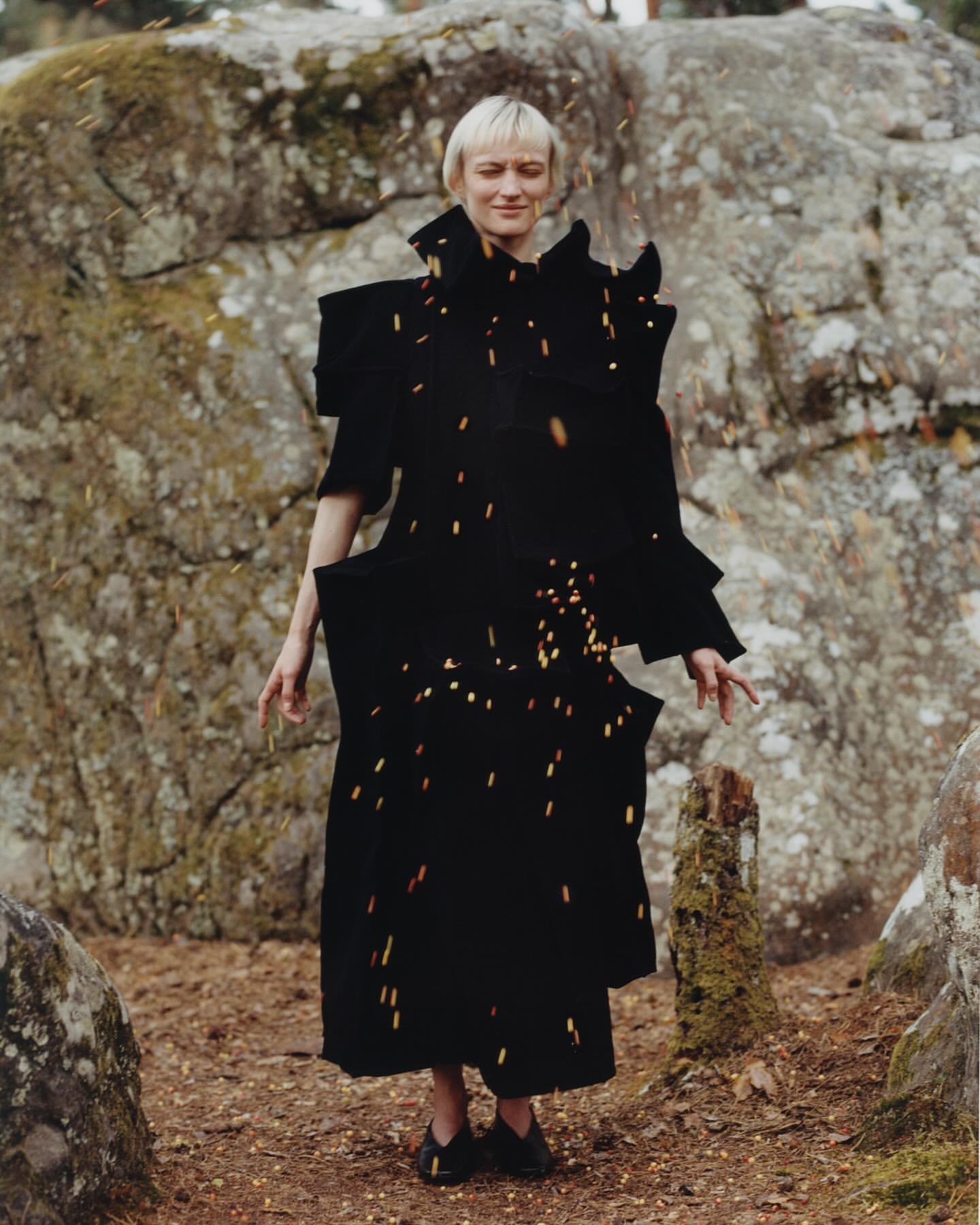 Female adult in conceptual fashion posing in a black coat, outdoors among rocks and falling debris in a medium shot in nature.