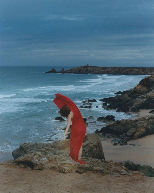 Woman in a red dress dances on a rocky cliffside overlooking a beach and ocean landscape with a dramatic sky above.
