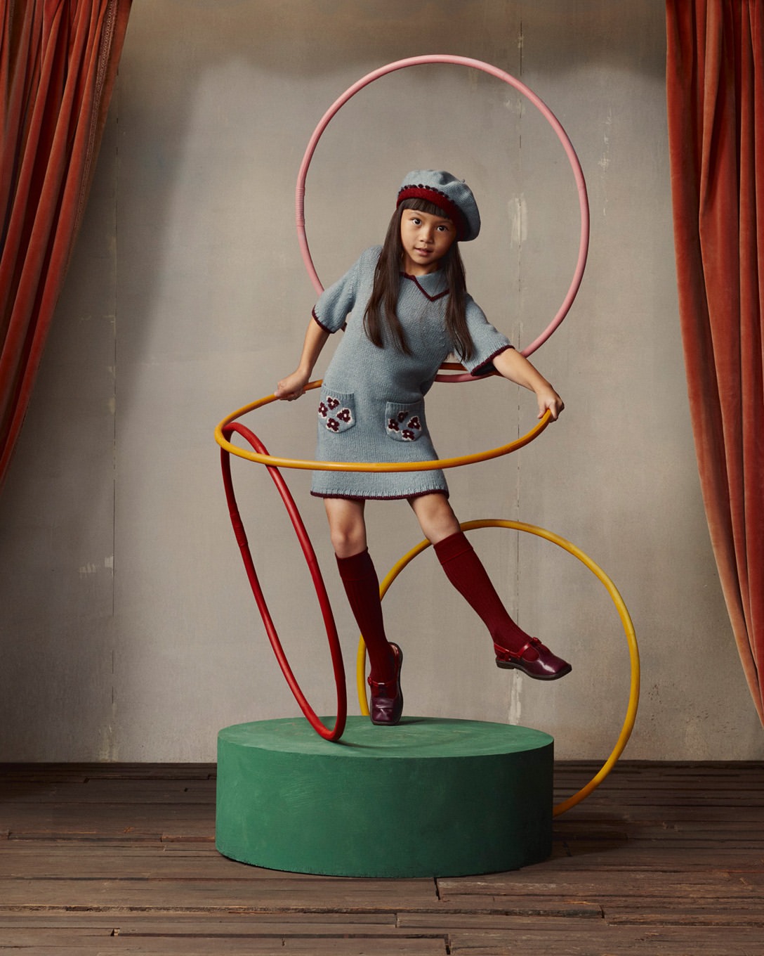 Young girl in a light blue dress and beret poses with hula hoops in a studio setting, standing on a green platform with curtains behind.