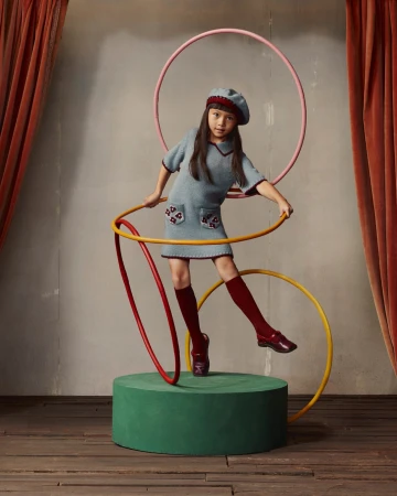 Young girl in a light blue dress and beret poses with hula hoops in a studio setting, standing on a green platform with curtains behind.