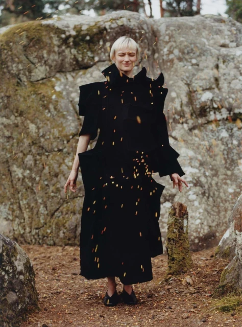 Adult female model stands in forest wearing dramatic black avant-garde dress. Brown forest floor, rock formations, and trees surround her.