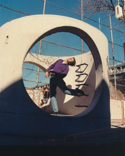 Adult male in purple sweater leans back inside of large concrete tunnel with graffiti art against a chain link fence under bright daylight.