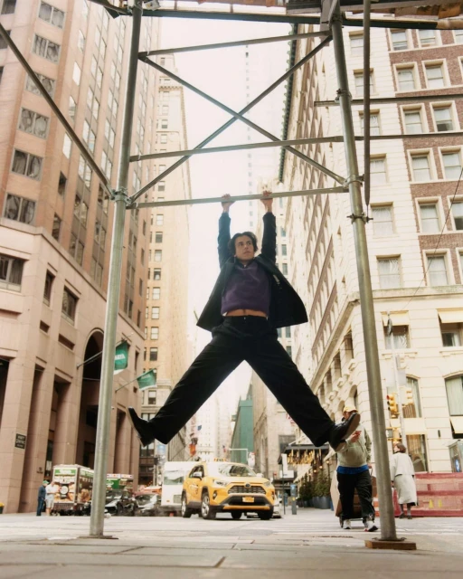 Man in business suit hanging from metal scaffolding on a city street with taxi, buildings, and pedestrian in the background.