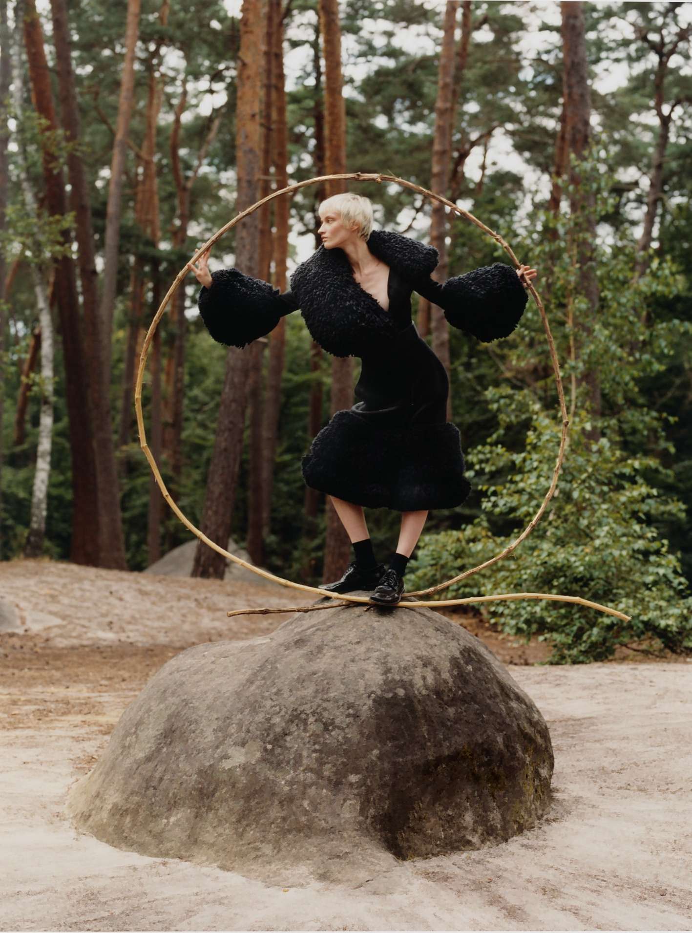 A woman in a black dress and boots standing on a rock in a forest holds a large hula hoop, creating a visually striking and unconventional pose.