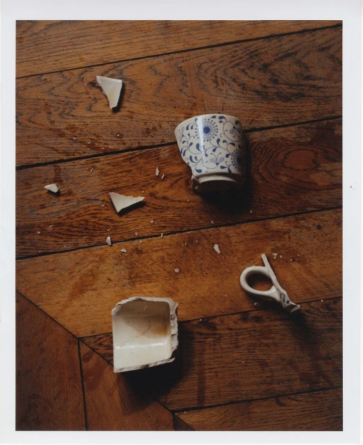 Still life of a shattered porcelain teacup with a blue floral pattern and broken white handle on a weathered hardwood floor surface.