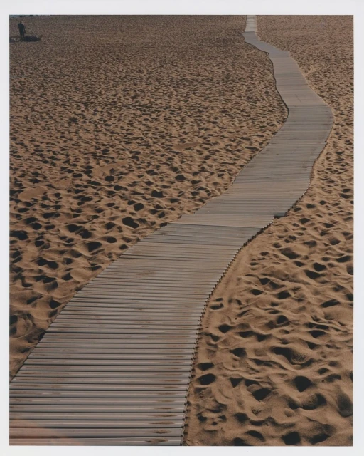 Wooden boardwalk curves through a sandy beach landscape, leading the eye through the scene with a solitary figure in the distance.