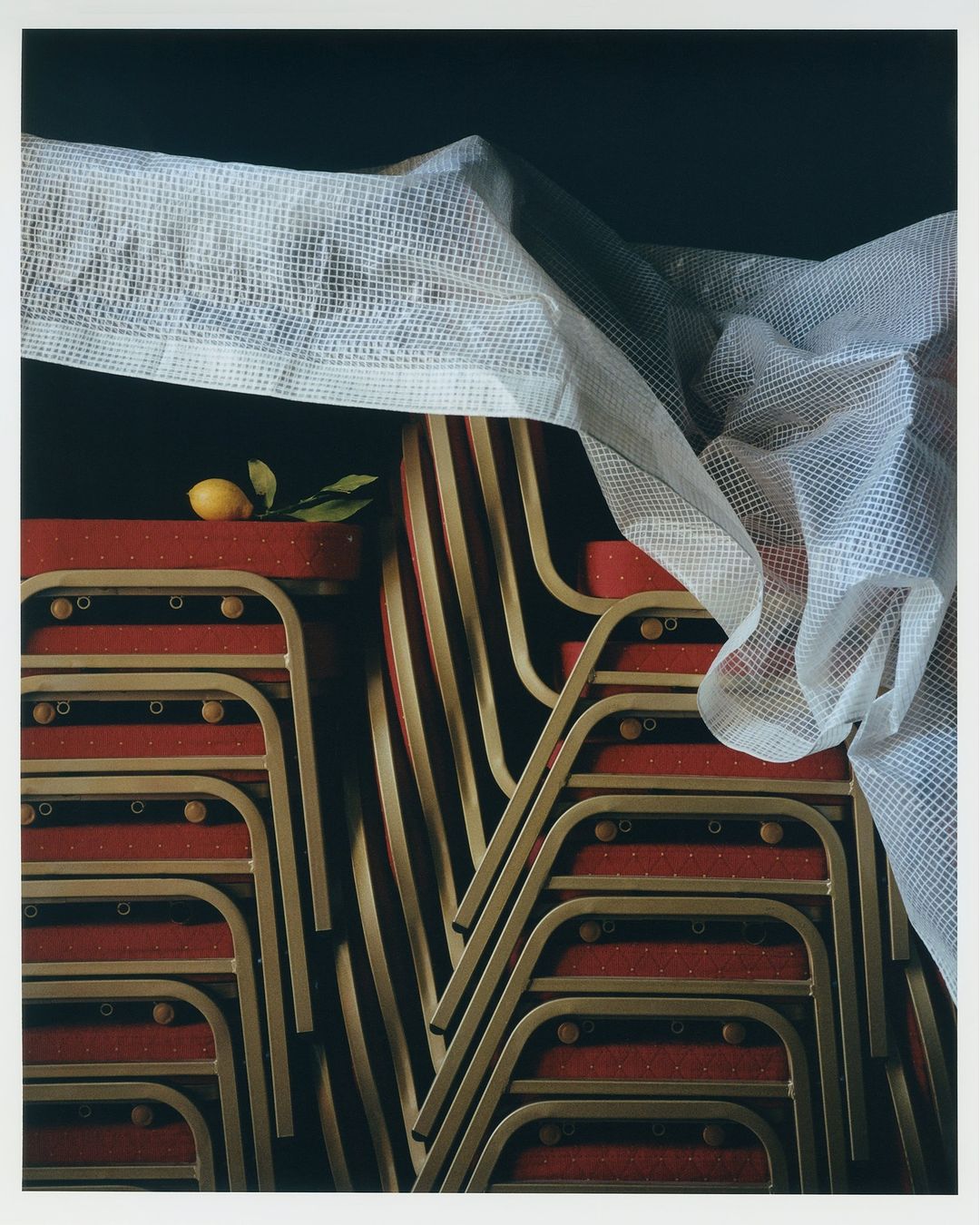 Still life photograph of stacked red and gold folding chairs covered with a white fabric and garnished with a single lemon and green leaves.