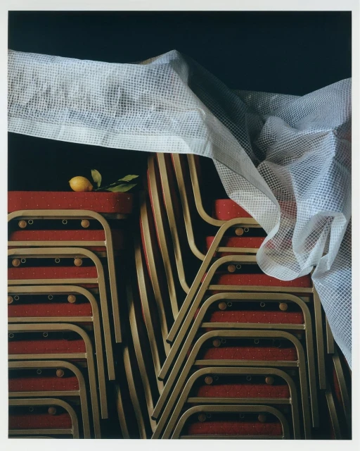 Still life photograph of stacked red and gold folding chairs covered with a white fabric and garnished with a single lemon and green leaves.