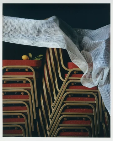 Still life photograph of stacked red and gold folding chairs covered with a white fabric and garnished with a single lemon and green leaves.
