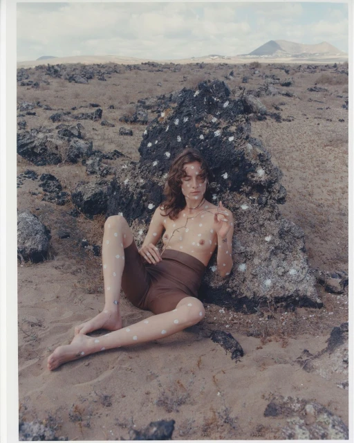 Bare-chested woman with white dots poses near a textured rock in a natural setting, wearing shorts and accessorized with a necklace.