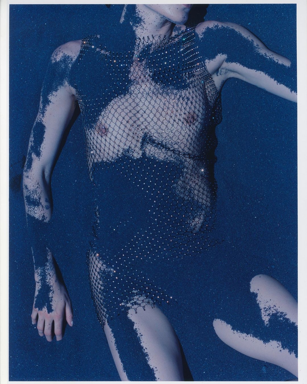 Fashionable adult woman laying down and covered in shimmering blue powder. She is wearing a sparkling net garment in a studio setting.