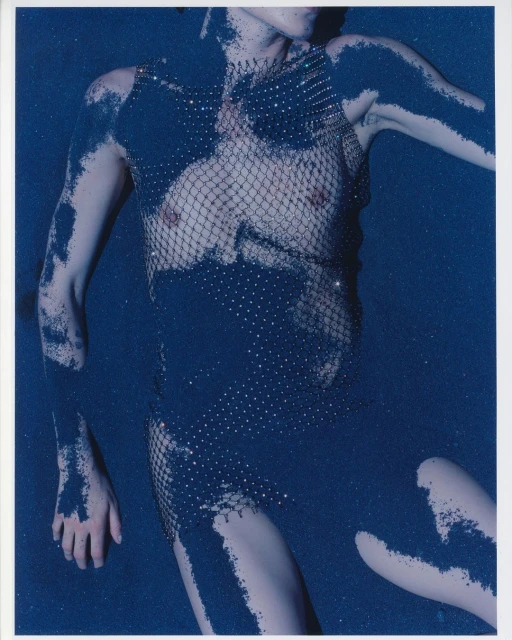 Fashionable adult woman laying down and covered in shimmering blue powder. She is wearing a sparkling net garment in a studio setting.