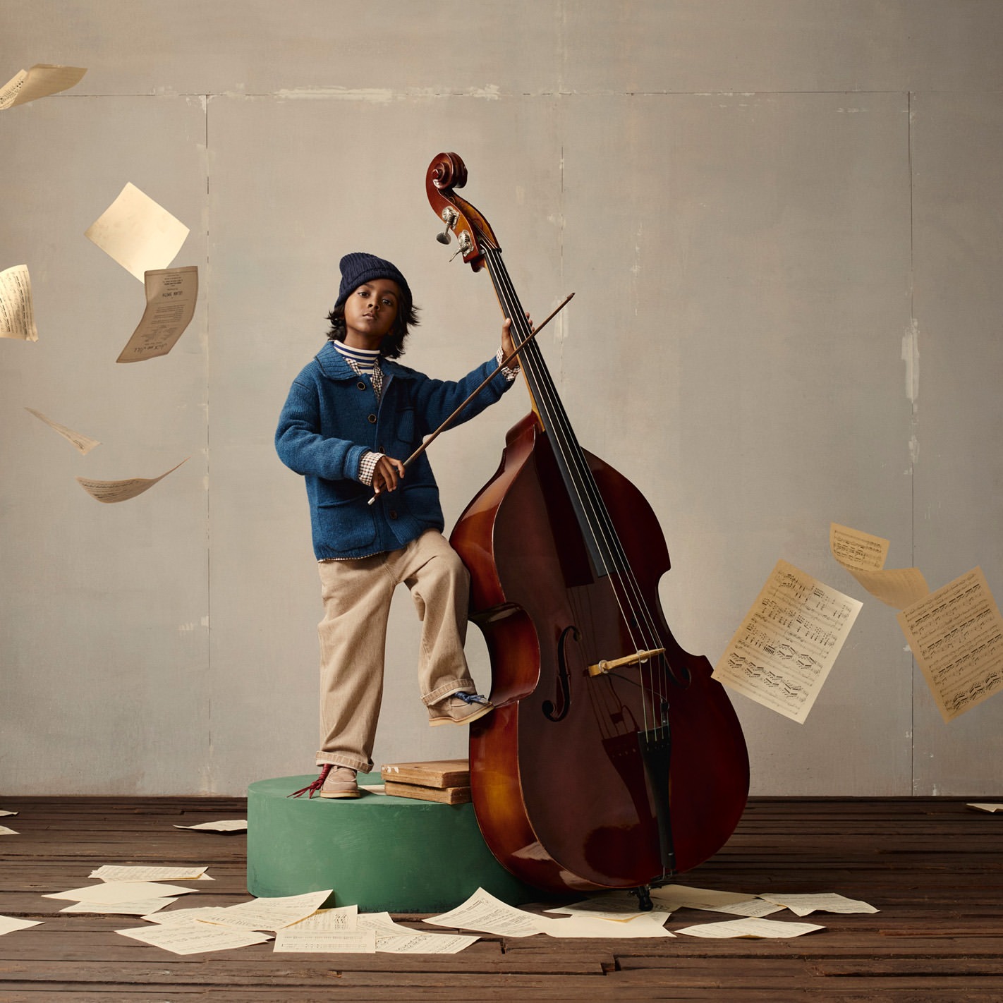 Young boy in a blue jacket stands near a large double bass in an indoor setting, sheet music scattered around, creating a musical atmosphere.
