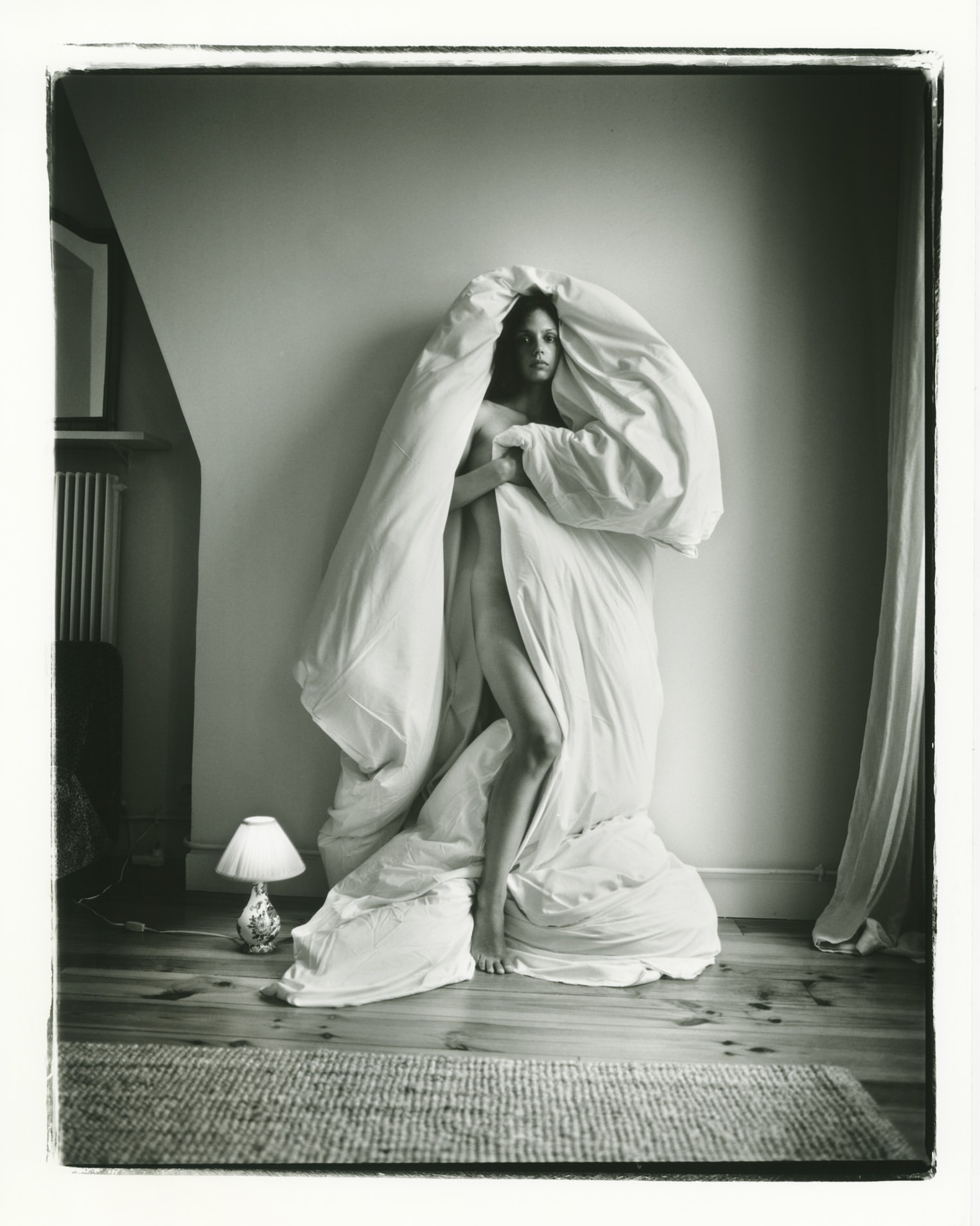 Monochrome portrait of a fair-skinned adult female standing indoors, draped in a white blanket or duvet, next to a small lamp.