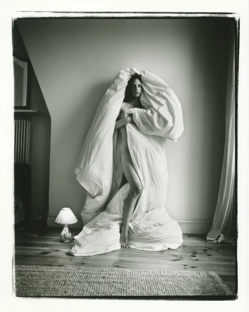 Monochrome portrait of a fair-skinned adult female standing indoors, draped in a white blanket or duvet, next to a small lamp.
