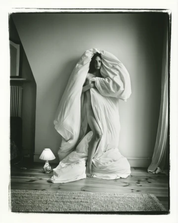 Monochrome portrait of a fair-skinned adult female standing indoors, draped in a white blanket or duvet, next to a small lamp.