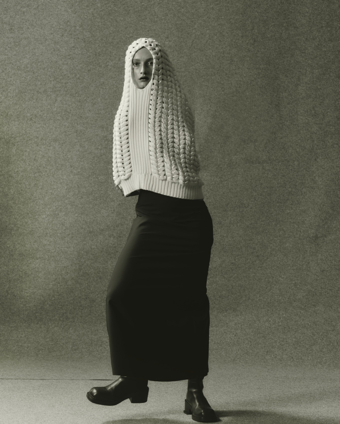 Full-length studio shot of a woman in a cream knit hooded top, long black skirt, and black boots against a neutral grey background.