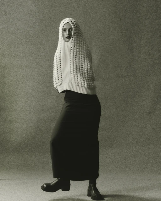Full-length studio shot of a woman in a cream knit hooded top, long black skirt, and black boots against a neutral grey background.