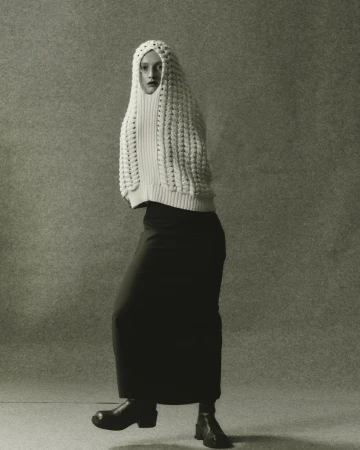 Full-length studio shot of a woman in a cream knit hooded top, long black skirt, and black boots against a neutral grey background.