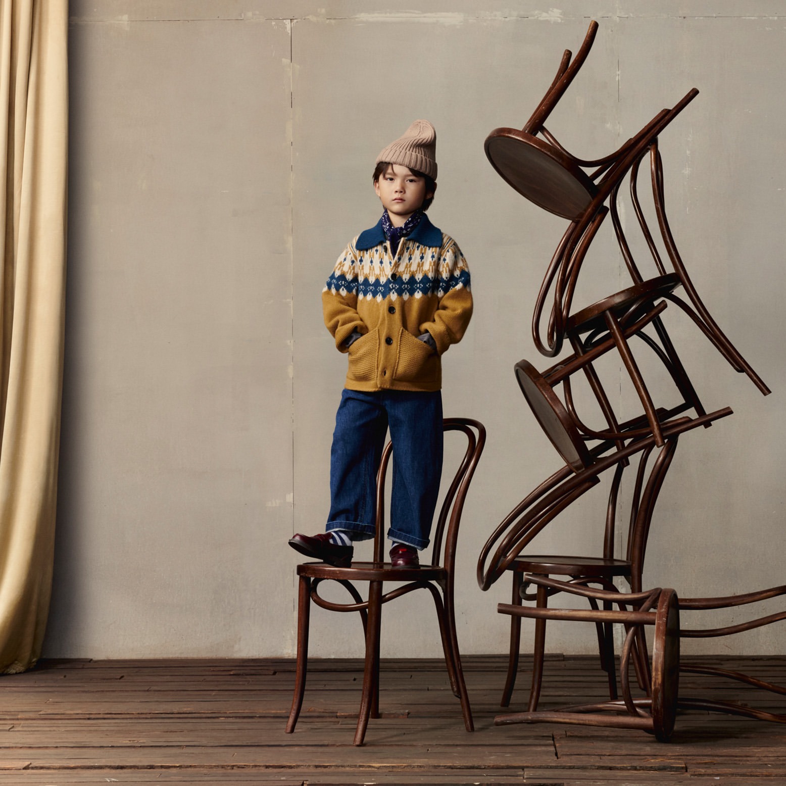 Young boy with a beanie and patterned sweater standing on a chair surrounded by a stack of bentwood chairs in a rustic indoor setting.