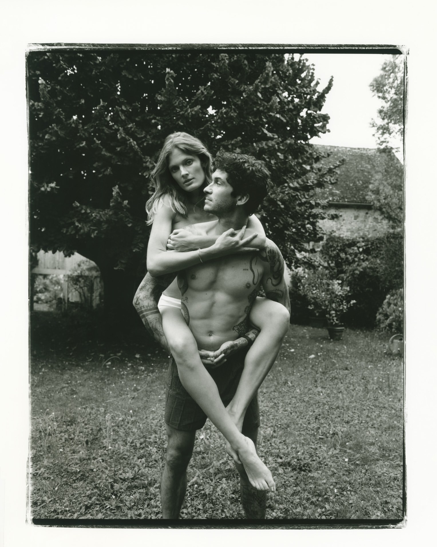 Black and white portrait of a woman being carried on a man back outdoors in a garden by Denis Boulze