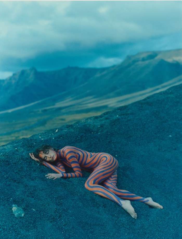 Woman with body paint lies on dark sand in an outdoor setting; mountains and cloudy sky in the background. Body art concept.