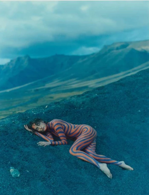 Woman with body paint lies on dark sand in an outdoor setting; mountains and cloudy sky in the background. Body art concept.