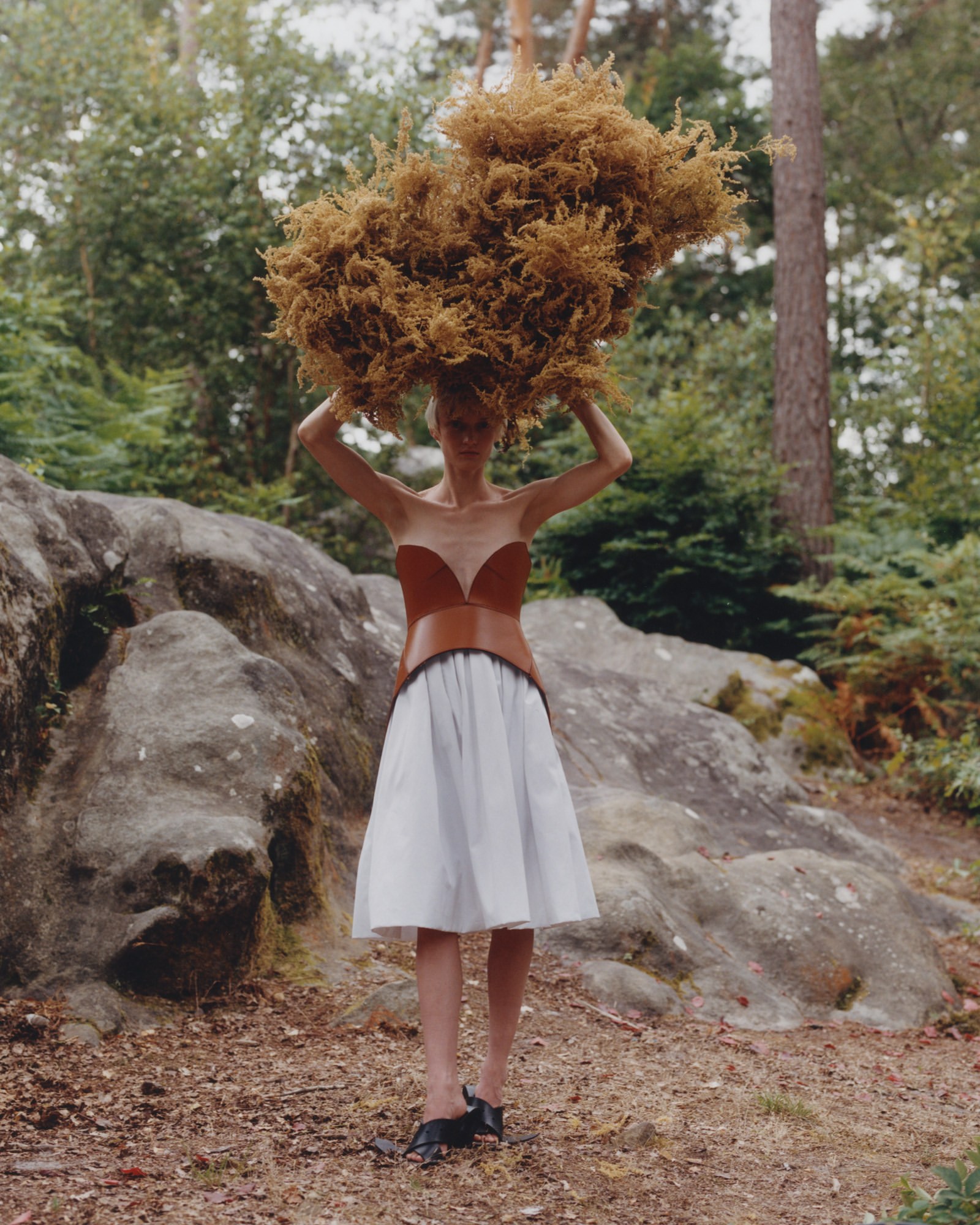 An adult woman in a sleeveless, strapless, rust-colored top and white skirt stands outside with an organic form atop her head.