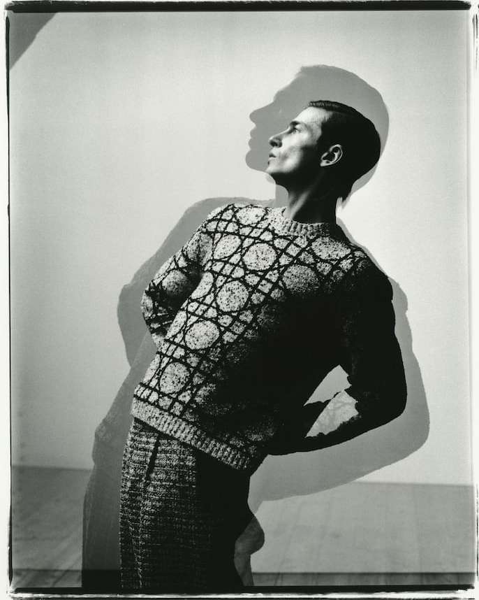 Monochrome portrait of a slender male model in patterned knitwear with hands behind his back, standing in front of a plain backdrop.