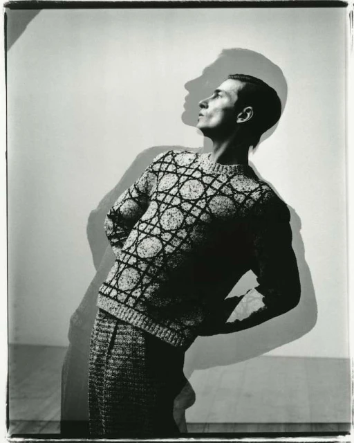 Monochrome portrait of a slender male model in patterned knitwear with hands behind his back, standing in front of a plain backdrop.