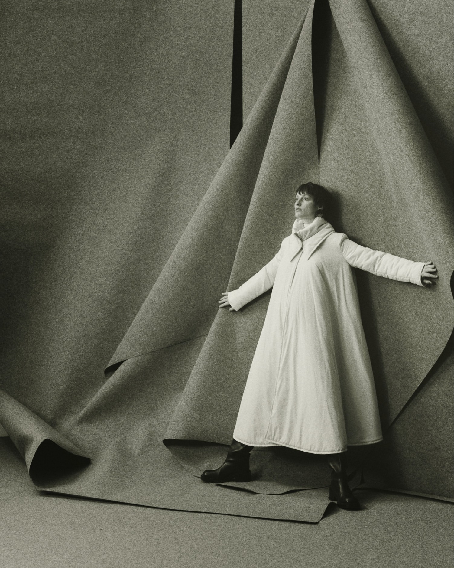 Monochrome fashion shot of an adult woman in a white coat, posing against folded gray fabric backdrop in a photo studio.